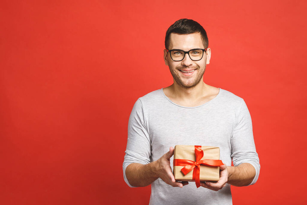 Wesołych świąt, moje kongregacje! Portret atrakcyjnego mężczyzny dającego pudełko prezentu i patrzącego na kamerę odizolowaną na czerwonym tle. - Zdjęcie, obraz