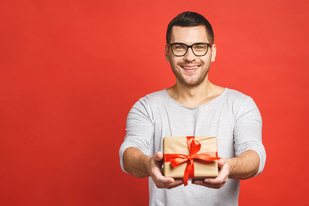 Happy holiday, my congredulations! Portrait of an attractive casual man giving present box and looking at camera isolated over red background. - Photo, Image