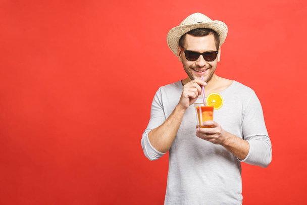 Portré vonzó fiatalember kalap és napszemüveg álló és ivás narancslé felett piros háttér. - Fotó, kép