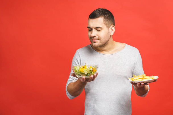 Портрет молодого красивого чоловіка, який прагне після нездорової їжі замість того, щоб їсти здорову їжу, ізольований на червоному тлі
.  - Фото, зображення