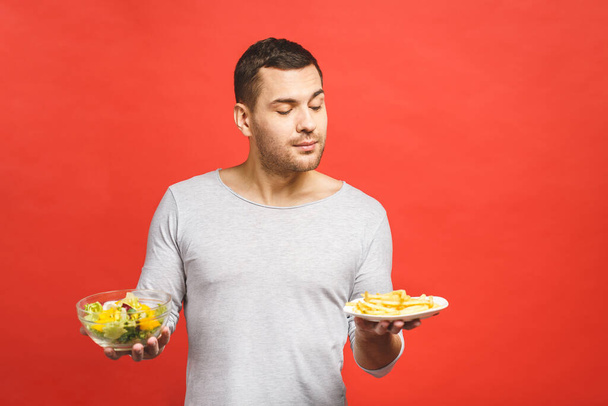Portrait of young handsome man craving after junk food instead of eating healthy food, isolated over red background.  - Photo, Image