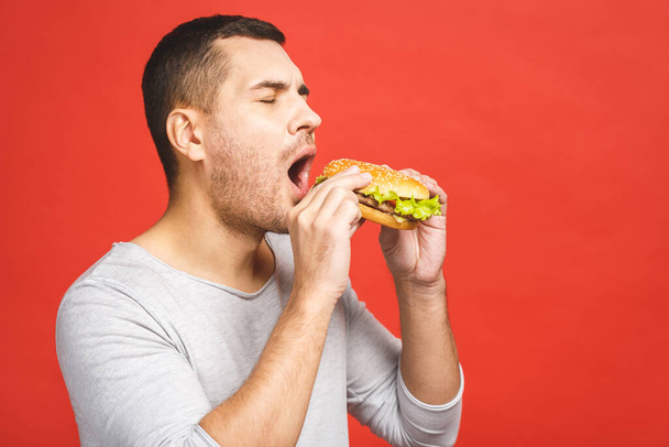 Young man holding a piece of hamburger. Student eats fast food. Burger is not helpful food. Very hungry guy. Diet concept. Isolated over red background. - Fotografie, Obrázek