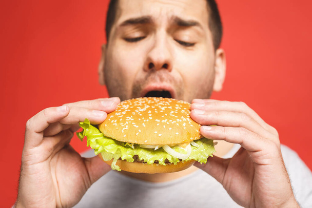 Young man holding a piece of hamburger. Student eats fast food. Burger is not helpful food. Very hungry guy. Diet concept. Isolated over red background. - Fotografie, Obrázek