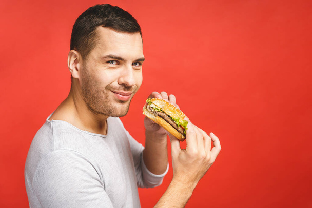 Mladý muž drží kus hamburgeru. Student jí fast food. Burger není užitečné jídlo. Velmi hladový chlap. Dietní koncept. Izolováno přes červené pozadí. - Fotografie, Obrázek