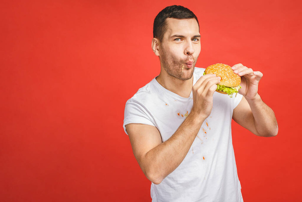 Vicces, éhes, szakállas ember gyorskaját eszik. Izgatott fiatalember mohón eszik hamburgert elszigetelve a vörös háttér. - Fotó, kép
