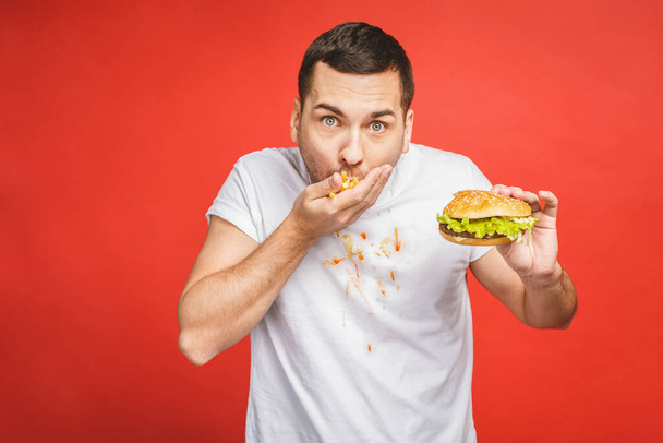 Divertido hombre barbudo hambriento comiendo comida chatarra. Joven emocionado comiendo hamburguesas aisladas sobre fondo rojo
. - Foto, Imagen