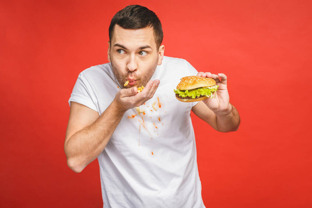 Смішний голодний бородатий чоловік їсть нездорову їжу. Захоплений молодий чоловік з жадібністю їсть гамбургери ізольовані на червоному тлі
. - Фото, зображення