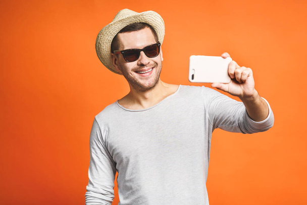 Cheerful young man in hat and sunglasses taking selfie with smartphone isolated over orange background. - Foto, Imagem