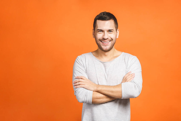 Casually handsome. Confident young handsome man keeping arms crossed and smiling while standing against orange background. - Photo, Image
