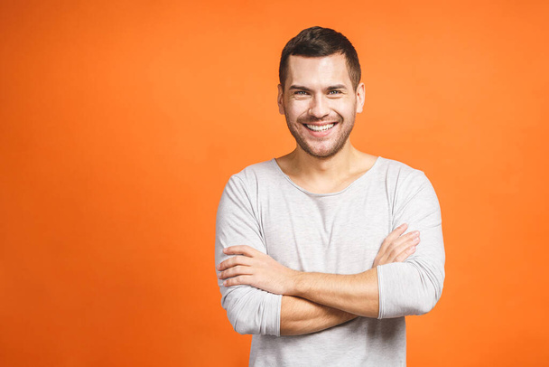 Casually handsome. Confident young handsome man keeping arms crossed and smiling while standing against orange background. - Foto, imagen