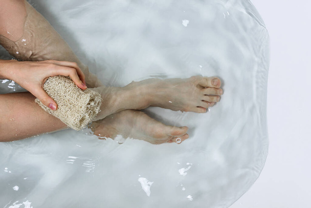 Above shot of woman's legs and hands holding luffa sponge in bathtub with clean water, bodypositive of self care concept - Fotografie, Obrázek