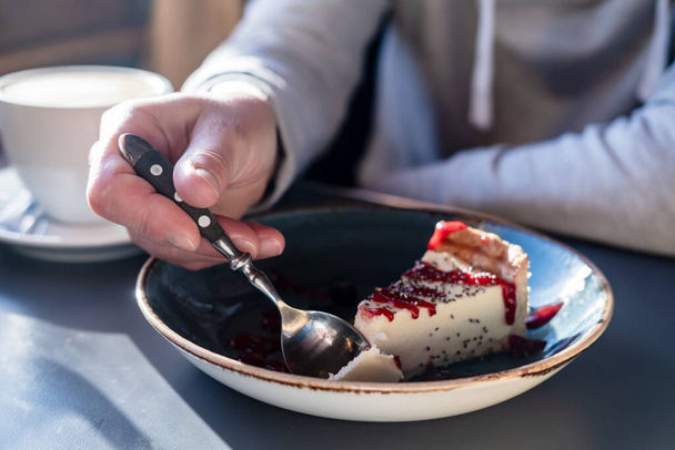 Lahodný tvarohový koláč se láme lžičkou z velkého kusu na krásném talíři, na rozmazaném pozadí šálek cappuccina. - Fotografie, Obrázek