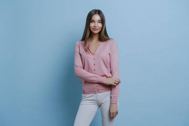 Fashionable brunette Caucasian woman dressed in pink jumper and white trousers, going to work, applies makeup, smiles pleasantly, isolated on blue background. People, style, lifestyle concept - Valokuva, kuva