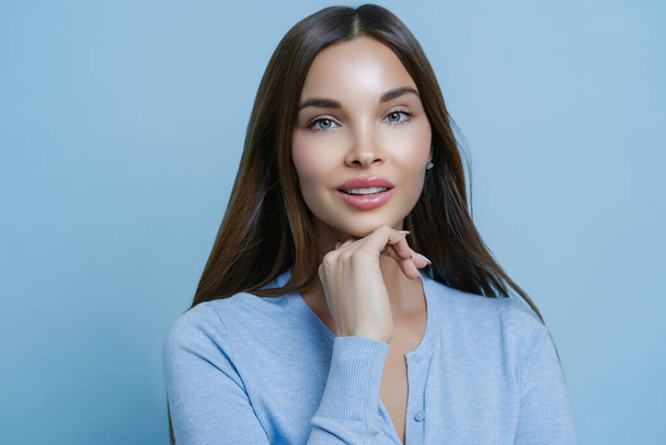 Beauty is in natural look. Portrait of pleasant looking European woman keeps hand under chin, looks with calm expression at camera, consults beautician, isolated over blue studio background. - Photo, Image