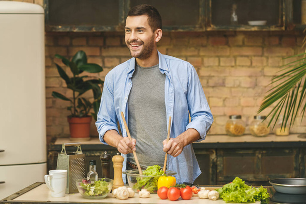 It's so delicious! Casual happy young man preparing salad at home in loft kitchen and smiling. - Photo, Image