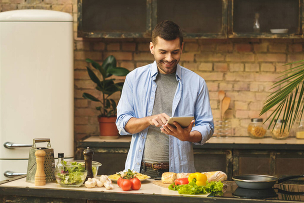 Adam güneşli bir günde ev mutfağında lezzetli ve sağlıklı yemekler hazırlıyor. Tablet bilgisayarı kullanılıyor. - Fotoğraf, Görsel