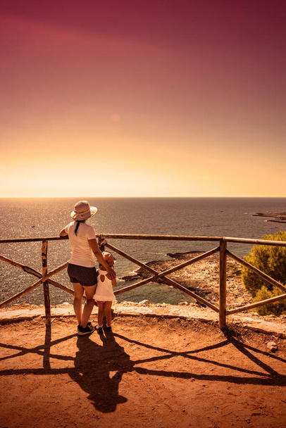 Bir anne ve küçük kızı manzaraya ve deniz manzarasına yukarıdan, panoramik bir terasta hayran kalırlar. Yazın Porto Selvaggio, Salento, Lecce, Puglia, İtalya 'da. Gün batımında. - Fotoğraf, Görsel