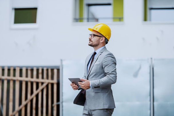 Вид збоку серйозного працьовитого архітектора в сірому костюмі і з жовтим шоломом на голові тримає планшет і дивиться на будівлю, стоячи на будівельному майданчику
. - Фото, зображення