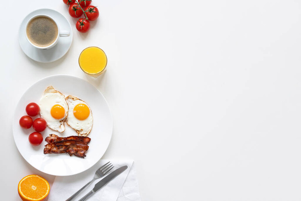 American breakfast with two eggs, bacon, tomatoes, orange, coffee and orange juice on white table. Top view. Copy space. Horizontal. - Photo, image