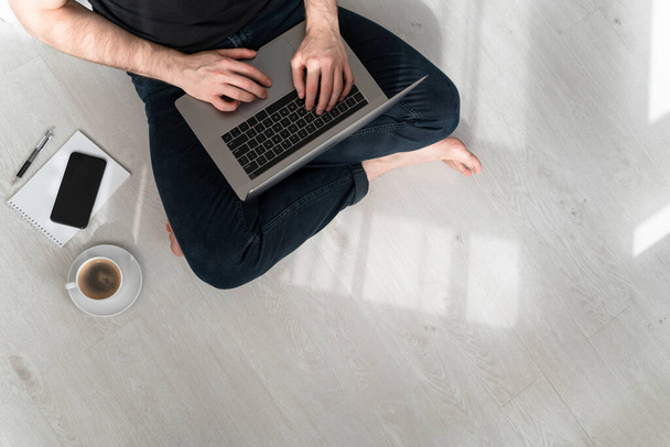 Man sitting on the floor at home with laptop, notebook, pen and cup of coffee. Online office. Work from home. Copy space.  Horizontal. - Valokuva, kuva