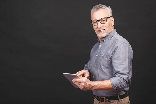 Great news! Handsome mature businessman in smart casual wear and eyeglasses is using a digital tablet and smiling, isolated against black background. Looking at camera.  - 写真・画像