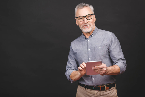 Great news! Handsome mature businessman in smart casual wear and eyeglasses is using a digital tablet and smiling, isolated against black background. - Photo, Image