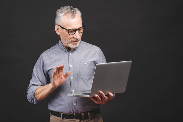 Χρησιμοποιώντας skype. Ηλικιωμένος γενειοφόρος γέρος με γυαλιά που κρατάει φορητό υπολογιστή απομονωμένο σε μαύρο φόντο. - Φωτογραφία, εικόνα