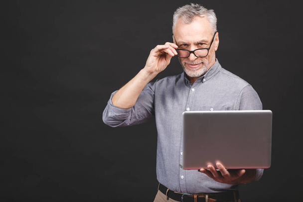 Senior aged bearded old man in eyeglasses holding laptop computer and smiling isolated against black background. - Photo, Image
