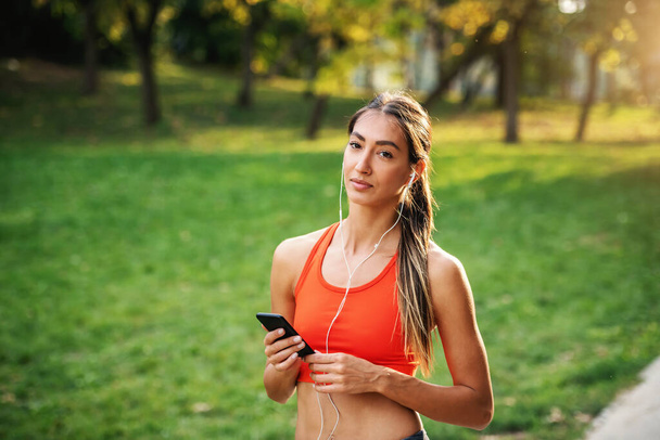 Young attractive pensive slim sportswoman standing in nature, having earphones in ears, holding smart phone and taking a break form running. - Photo, image