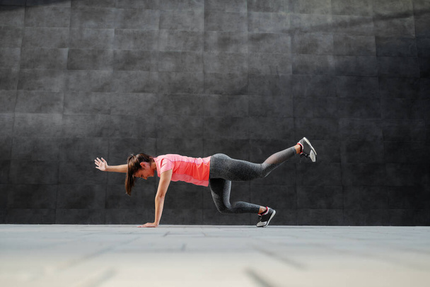 Side view of smiling attractive caucasian brunette in sports wear doing alt arm leg planks outdoors. In background is dark wall. - Foto, afbeelding