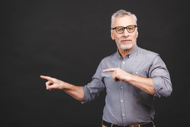 Aged senior man wearing glasses isolated against black background amazed and smiling to the camera while presenting with hand and pointing with finger. - Photo, Image