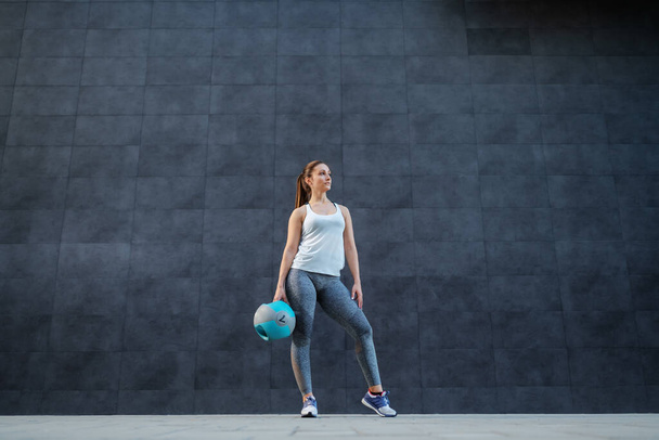手とポージングで体重ボールで外に立ってスポーツウェアで魅力的なフィット白人のスポーツウェアの女性。背景は暗い壁。. - 写真・画像