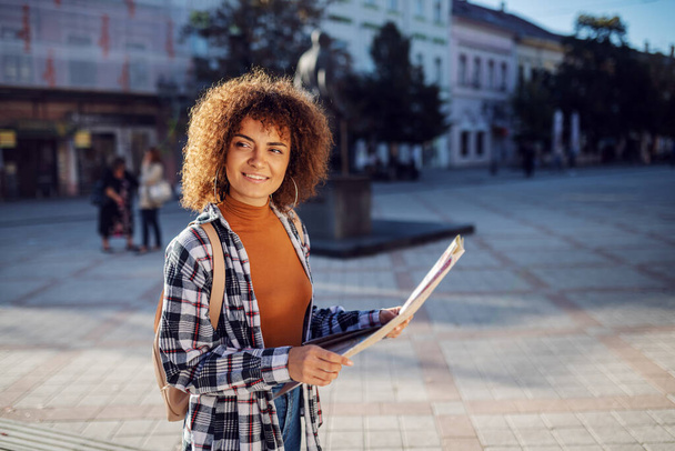 Молодая привлекательная женщина-туристка стоит на площади, держит карту и смотрит вокруг
. - Фото, изображение