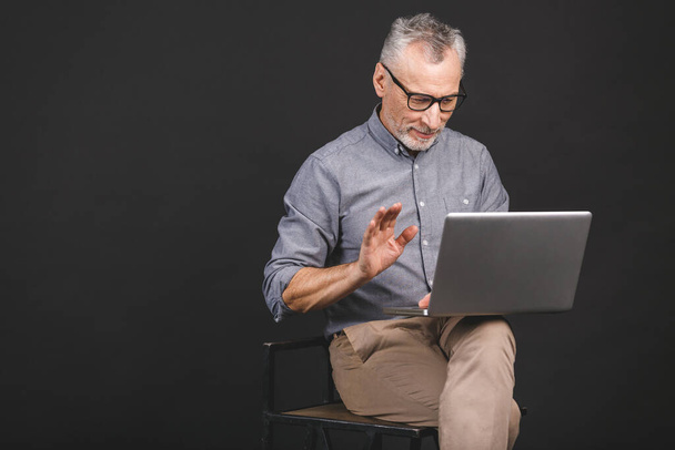 Дедушка использует современные технологии. Улыбающийся старый бизнесмен в очках, использующий ноутбук, изолированный на черном фоне
. - Фото, изображение