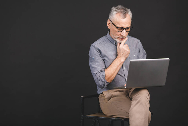 Дедушка использует современные технологии. Улыбающийся старый бизнесмен в очках, использующий ноутбук, изолированный на черном фоне
. - Фото, изображение