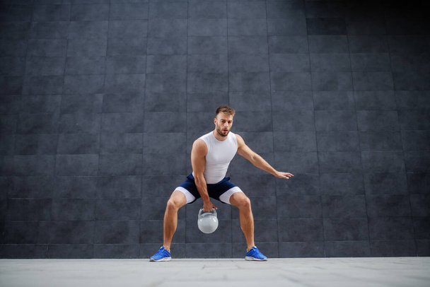 Strong muscular handsome Caucasian man in shorts and t-shirt standing outdoors and swinging kettle bell. In background is gray wall. - Zdjęcie, obraz