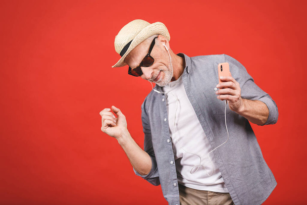 Retrato de un anciano alegre y mayor bailando y escuchando música con teléfono y auriculares aislados sobre fondo rojo
. - Foto, imagen