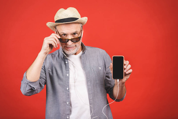 Весёлый пожилой человек слушает музыку на телефоне, изолированном от красного фона
. - Фото, изображение