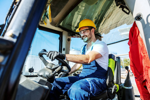 Smiling handsome caucasian worker in overall and with helmet on head driving excavator. - Fotó, kép