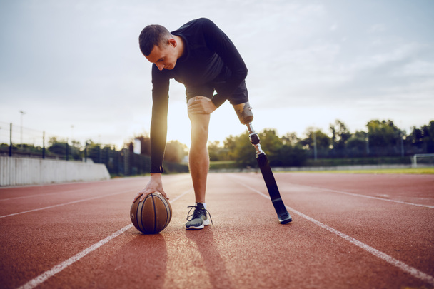 Pełna długość sportowych kaukaski niepełnosprawny mężczyzna w odzieży sportowej i sztuczne nogi dryblować piłkę stojąc na torze wyścigowym. - Zdjęcie, obraz