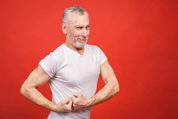 Check this out! Senior man showing muscle. Aged man flexing his arms isolated against red background. Cheerful excited modern cool pensioner grandpa practising bodybuilding. - Zdjęcie, obraz