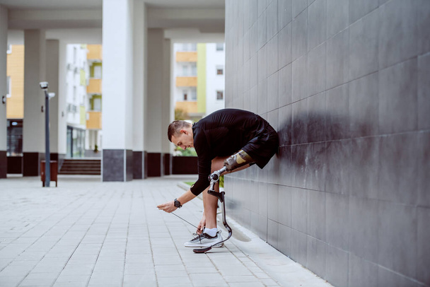 Side view of attractive caucasian sportsman with artificial leg leaning on the wall and tying his shoelace. - Fotó, kép