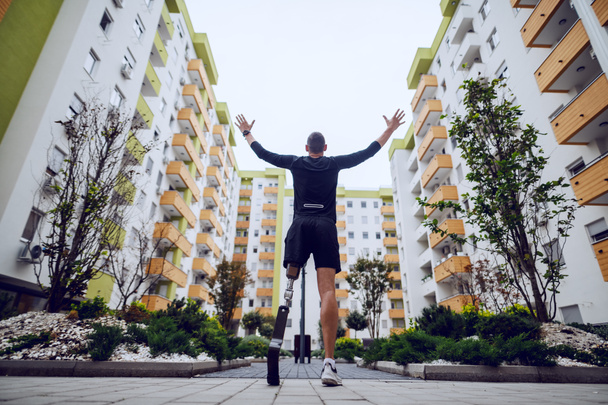 Rear view of handsome sportsman with artificial leg standing with hands in the air outdoors surrounded by buildings. - Photo, Image