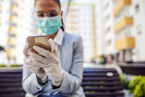 Young gorgeous businesswoman with protective mask and gloves typing message on smart phone while sitting in park after work. Selective focus on phone. - Foto, immagini