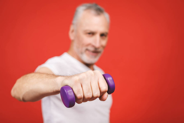 Közelkép portré egy idős ember gyakorlása súlyzókkal szemben piros háttér. - Fotó, kép