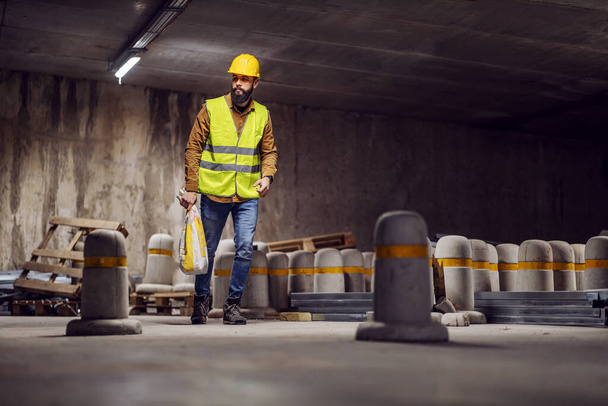 Longitud completa de la bolsa de transporte de trabajadores con cemento mientras camina en el garaje subterráneo en proceso de construcción
. - Foto, Imagen