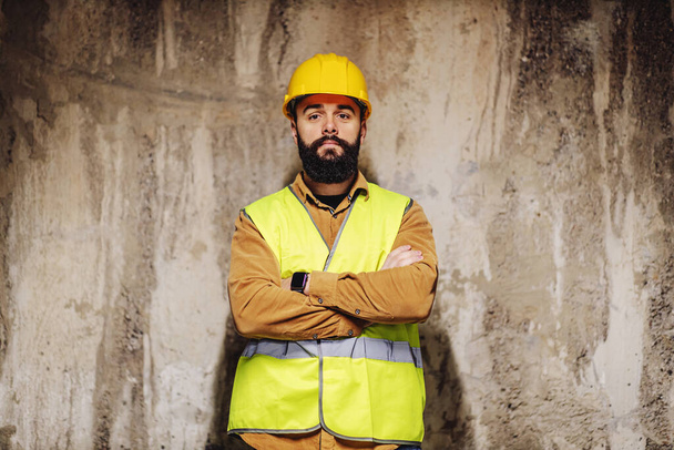 Fiatal szakállas munkás áll az építkezésen keresztbe tett karokkal. - Fotó, kép