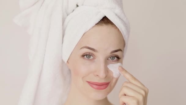 Portrait of charming Caucasian woman in white towel, applying white clay beauty mask on her face with her finger. Spa, beauty procedures at home, healthcare. - Imágenes, Vídeo