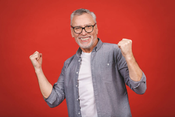 Happy mature senior man in casual using smartphone and making winner gesture isolated over red background. - Photo, Image