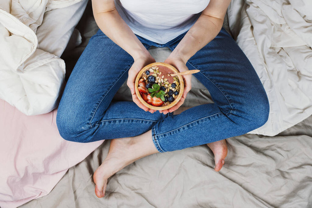 Top view of woman in jeans and white shirt in her bed holding vegan smoothie bowl, selective focus - Φωτογραφία, εικόνα